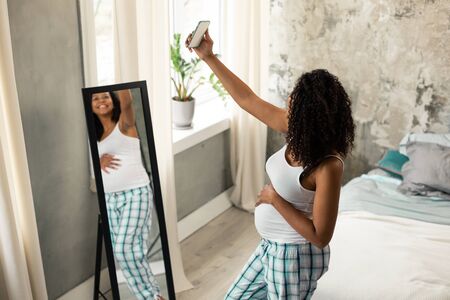 Taking a picture. Happy smiling pregnant woman standing in front of her mirror at home and taking a selfie.の写真素材