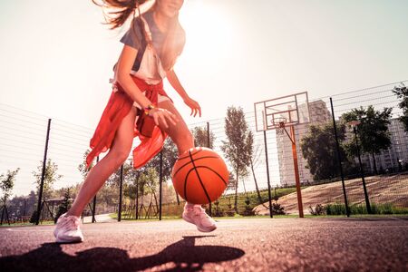 Sunny evening. Experienced dark-haired young girl running around basketball playground with orange ballの写真素材
