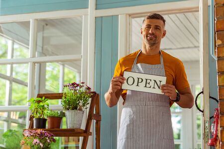 Ready to work. Smiling handsome man holding an open sign and standing near the doorway of his flower shop.の写真素材