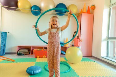 Playful child. Delighted blonde girl smiling while playing with a hoop