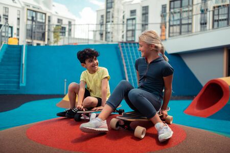 Making new friends. Two cheerful children talking sitting on their skateboards outside.