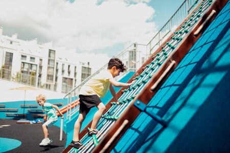 Outdoor activities. Concentrated schoolboy climbing a rope ladder with his classmate on a sports...