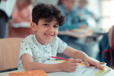 Happy mood. Cheerful pupil sitting at his school desk, writing a test and happily smiling.の写真素材