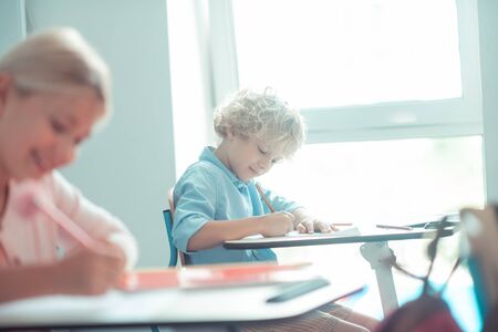 Writing the test. Smiling blond-haired boy writing his test sitting near the window in the class.の写真素材