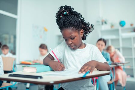 Solving problems. Cute schoolgirl sitting in the classroom and doing sums with concentrated face at the test.の写真素材
