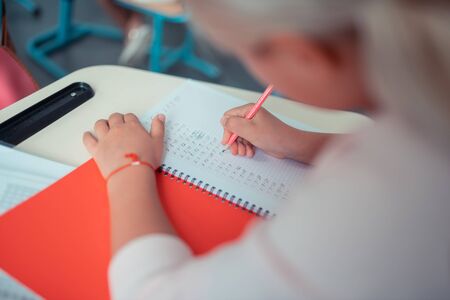 Loving maths. Concentrated schoolgirl doing sums in her red copybook sitting at the desk.