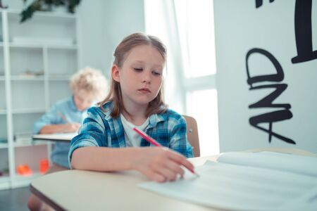 Waiting for the end of the lesson. Tired little school girl with a red pencil writing her maths test in the class.の写真素材