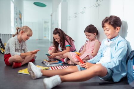 Addicted to smartphones. Stylish good-looking children addicted to smartphones using them during break at schoolの写真素材