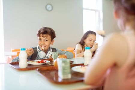 Lunch with classmates. Dark-haired boy smiling while having lunch with classmates during school breakの写真素材