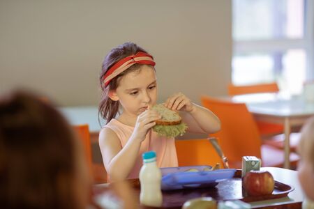 Eating yummy sandwich. Cute dark-eyed girl eating yummy sandwich with lettuce in canteenの写真素材