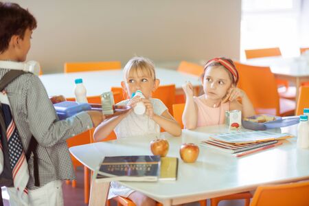 Boy joining girls. Dark-haired boy holding tray with food joining classmates in the school canteenの写真素材