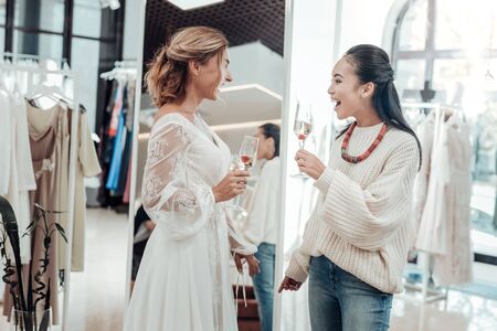 Cheerful mind. Beautiful woman wearing wedding dress and her smiling bridesmade drinking chamoagne in a wedding salon.の写真素材