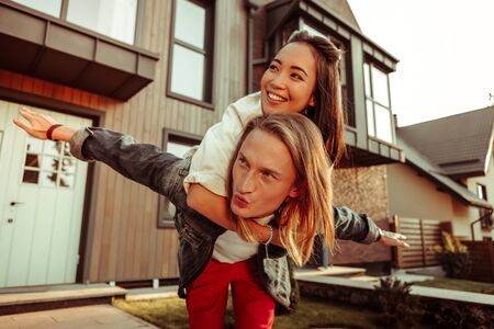 Having building behind. Active long-haired man in red pants spreading hands while carrying girl and entertaining herの写真素材