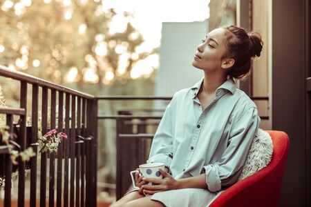 Drinking morning coffee. Peaceful good-looking woman in oversize male shirt carrying big cup while staying on a terraceの写真素材