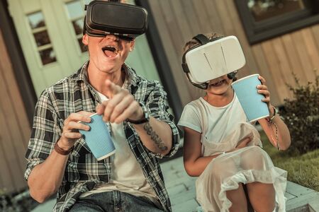 Wearing VR-helmets. Laughing active father in black and white shirt playing in VR-helmet with cheerful daughterの写真素材