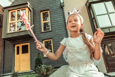Wearing mascaraed outfit. Positive long-haired girl in festive dress playing in fantastic game while sitting on the porchの写真素材