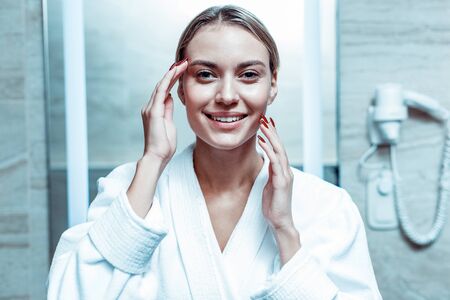 Wearing white robe. Cheerful good-looking lady with tied hair enjoying her flawless skin while staying in a bathroomの写真素材