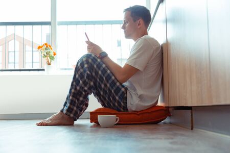 Sitting on floor. Handsome young man wearing pajamas sitting on the floor and using smartphone