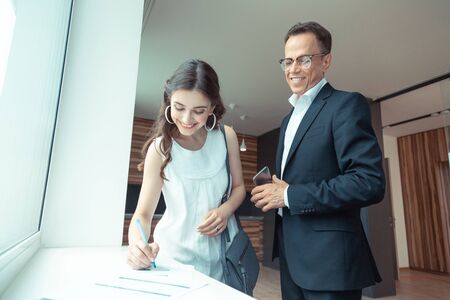 Client signing papers. Real estate agent wearing glasses watching the client signing the papers after buying houseの写真素材