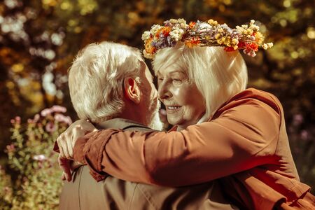Life partners. Thoughtful happy senior woman wearing flower wreath in arms of her husband in a park.の写真素材
