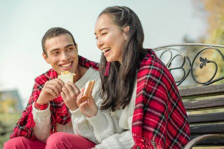 Joyful good-looking couple eating freshly-made sandwiches on a bench during lovely dateの写真素材