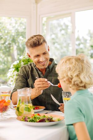 Handsome caring husband giving some tomato for his son while having lunchの写真素材