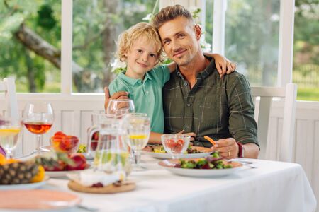 Dark-eyed handsome cute son hugging father while having yummy lunch