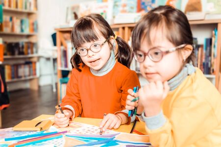 Creating drawings. Beaming little kids with down syndrome attentively looking while sitting in front of colorful papersの写真素材
