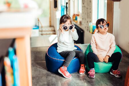 Unique look. Calm young children sitting together on the armchairs with sunglasses onの写真素材