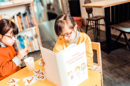 Words and alphabet . Focused dark-haired girl in yellow sweater attentively reading book about understanding of yourselfの写真素材