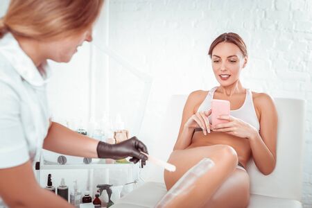 Dealing with wax. Pleasant young woman lying in white underwear while master coating her legs with special processed waxの写真素材