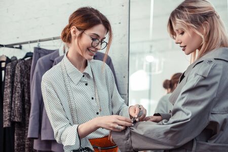 Choose trench coat. Pleasant fashion designer wearing glasses helping client to choose trench coatの写真素材