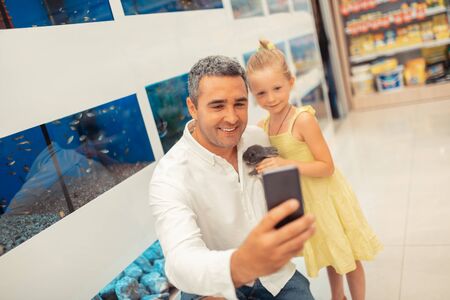 Selfie with daughter. Man making selfie with daughter while visiting pet shop and holding little...