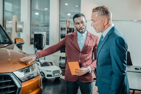 Giving advice. Enthusiastic car dealer standing near his interested client helping him buying a new car.の写真素材