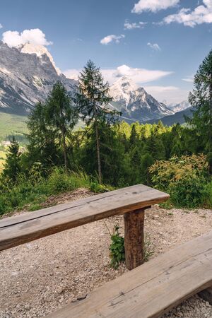 Beautiful landscape with high rocky mountain, green forest, wooden table and bench under blue skyの写真素材