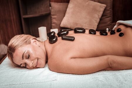 Feeling the warmth. Smiling beautiful woman lying on the towel in massage salon holding hot stones on her back.の写真素材