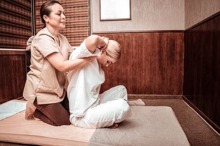 Body care. Smiling blond woman sitting on a mattress in front of masseuse holding her under hands and getting her thai massage in a salon.の写真素材