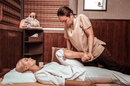 Taking care of body. Relaxed woman lying on the floor in a massage parlour getting her thai massage session by a strong masseuse bending her leg.の写真素材