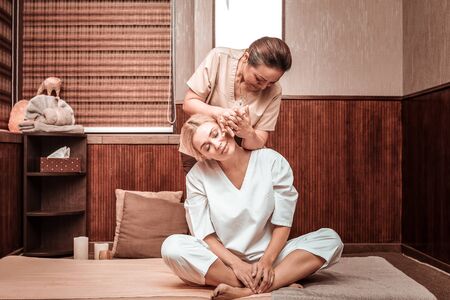 Gifted hands. Concentrated smiling masseuse doing neck and back massage for her happy client sitting on the mat.の写真素材