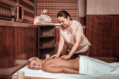 New experience. Cheerful beautiful woman lying on a mat in massage salon and getting her hot stone massage.の写真素材