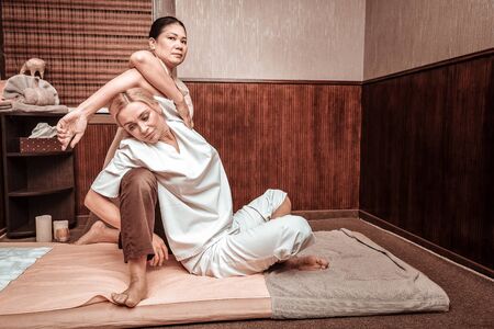 Working on strong body. Beautiful blond woman sitting crossing her legs and leaning over a knee of masseuse doing exercises during thai massage session.の写真素材