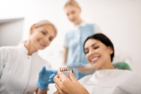 What suits better. Smiling woman choosing color for whitening her teeth sitting in the chair in dental clinic.の写真素材