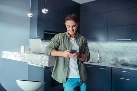 Man texting friend. Handsome young man standing in the kitchen and texting friendの写真素材