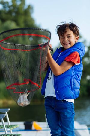 Net with fish. Happy beaming dark-haired boy holding fishing net with little fishの写真素材