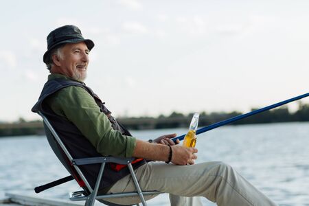 Enjoying evening. Retired man smiling while enjoying evening with fishing and drinking beerの写真素材