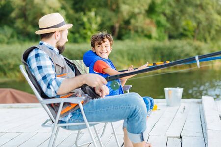 Looking at father. Cheerful son smiling and looking at father while fishing together near lakeの写真素材