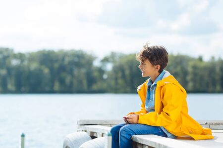 Cheerful boy. Cheerful boy wearing yellow raincoat sitting near river and breathing fresh airの写真素材