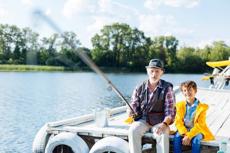 Male fishing. Bearded grey-haired pensioner wearing summer hat sitting near grandson and fishingの写真素材