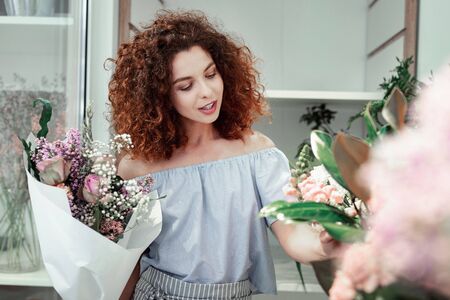 Different flowers. Interested curly-haired young lady touching stems of fresh flowers while choosing materials for her workの写真素材