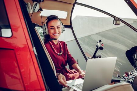 Before flying. Beautiful beaming businesswoman using laptop sitting in helicopter before flying